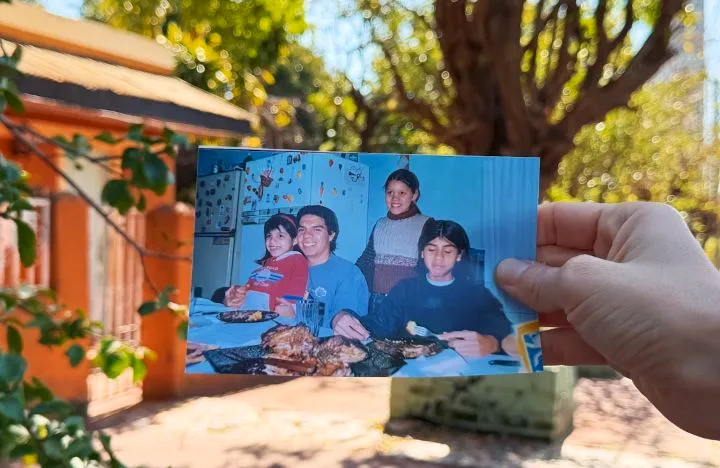 Una mano sostiene una foto familiar vieja frente a la casa naranja de la infancia, en una vereda de Posadas