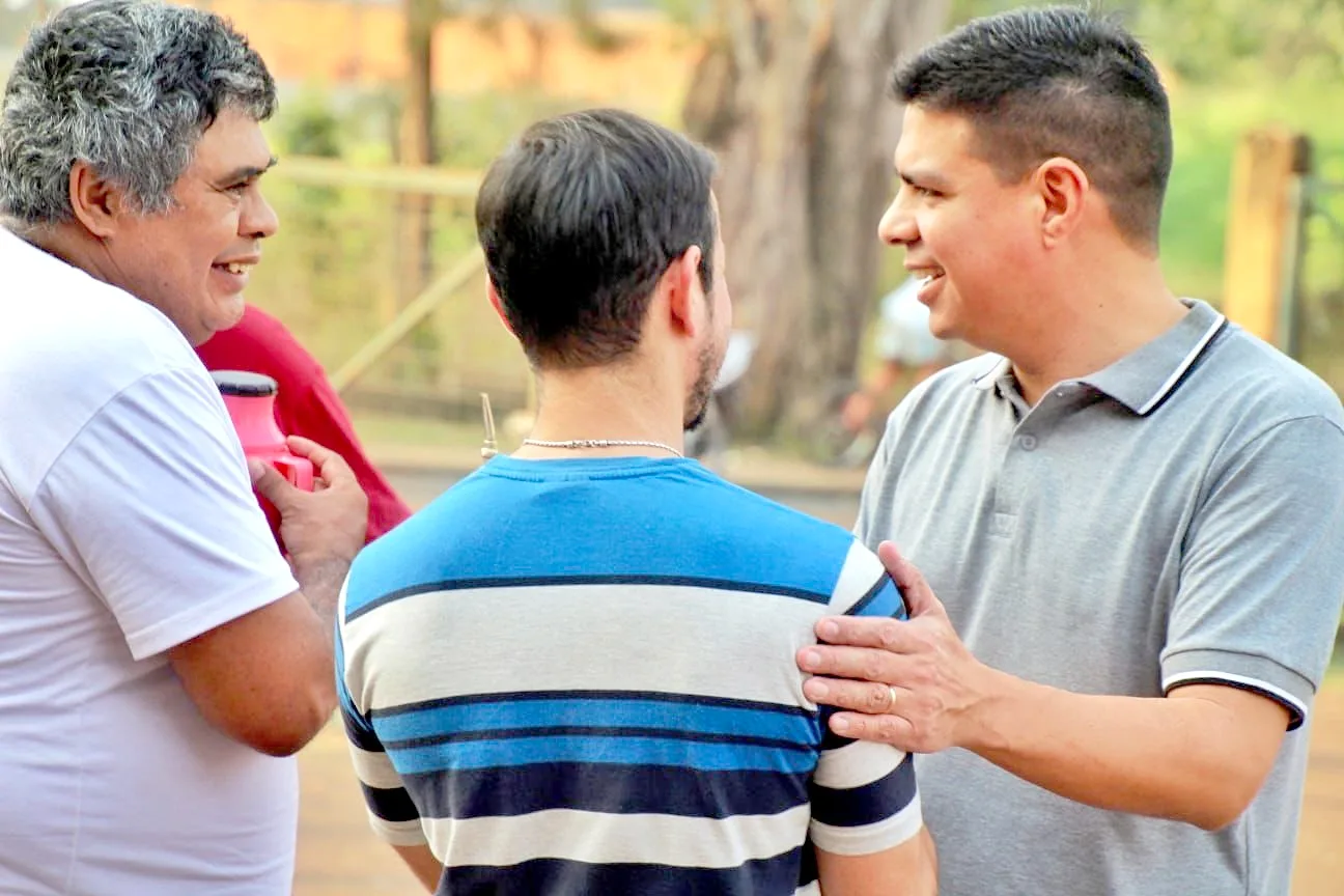 Fernando charla con dos vecinos bajo los árboles, con una mano en el hombro de uno de ellos, sobre tierra colorada