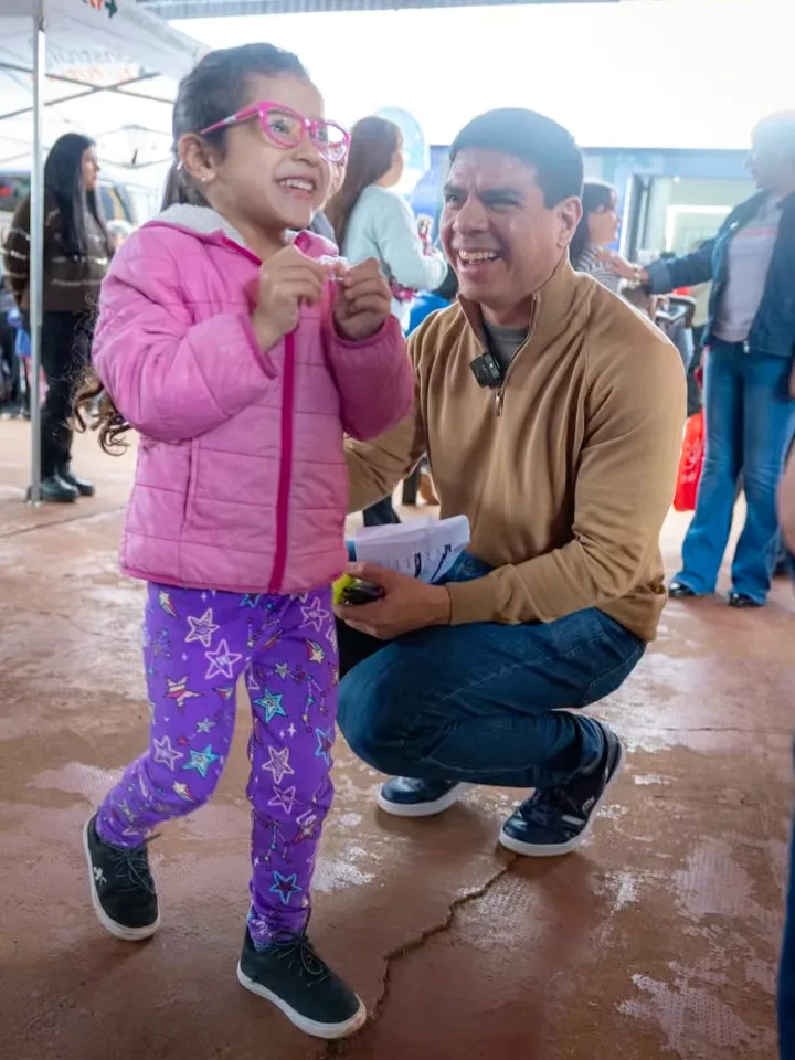 Fernando en cuclillas junto a una nena con lentes rosas nuevos, sobre la tierra colorada de un operativo en el barrio